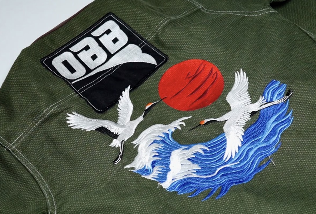 Embroidered white cranes flying over blue waves and a red sun on the back of the olive gi